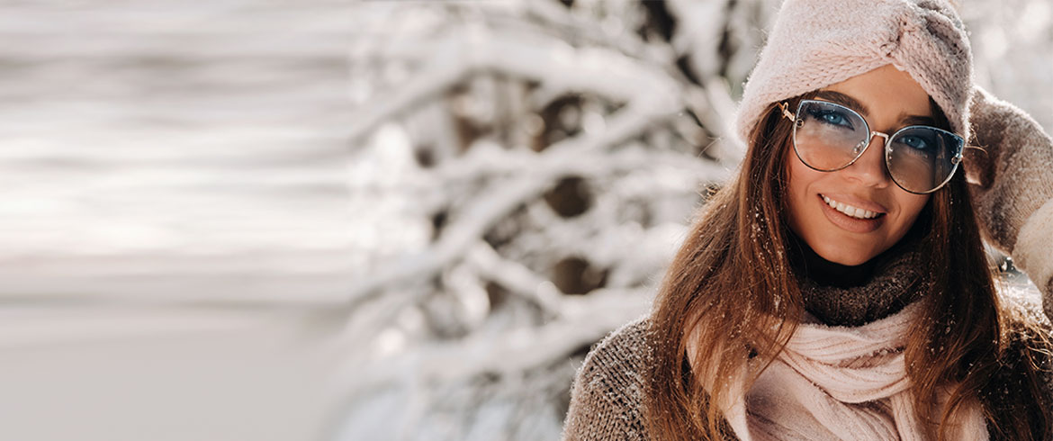 Eine lächelnde Frau mit Brille und warmer Kleidung genießt den Schnee, ihre Haut strahlt dank optimaler Winterpflege und Schutz vor Kälte.