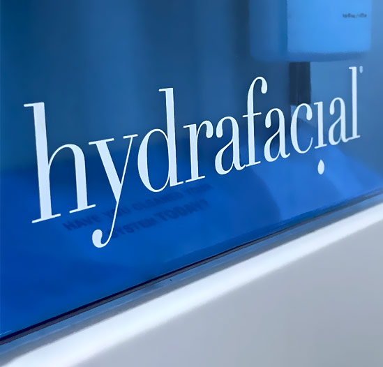 Detailaufnahme eines HydraFacial Geräts mit dem eingravierten Logo 'HydraFacial' auf der Oberfläche, umgeben von blauen Pflegeprodukten.